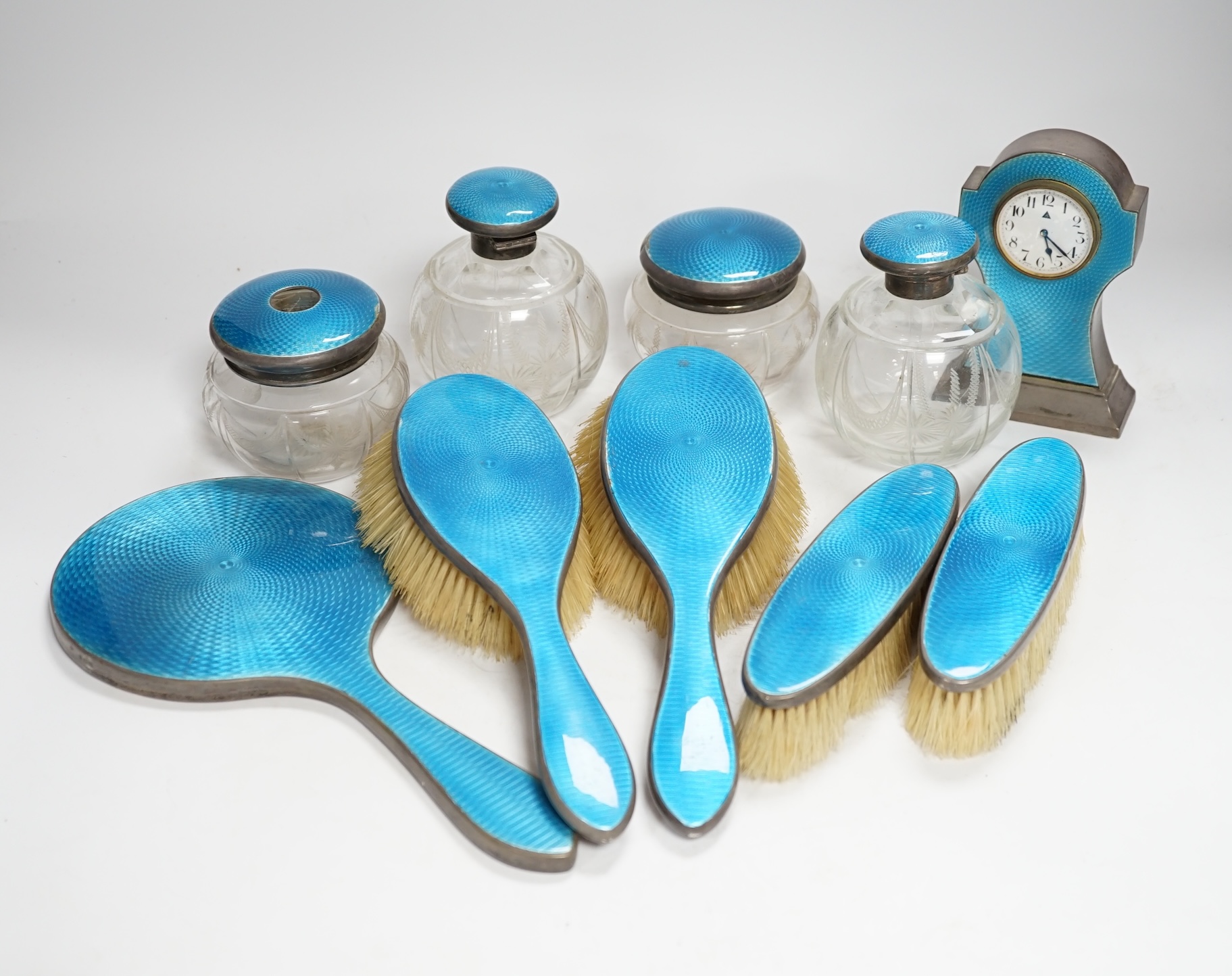 A George V silver and blue guilloche enamelled ten piece dressing table set, comprising four brushes, a hand mirror, two mounted glass toilet jars, two mounted glass scent and a mantel timepiece, 12.8cm, C.S Green & Co,